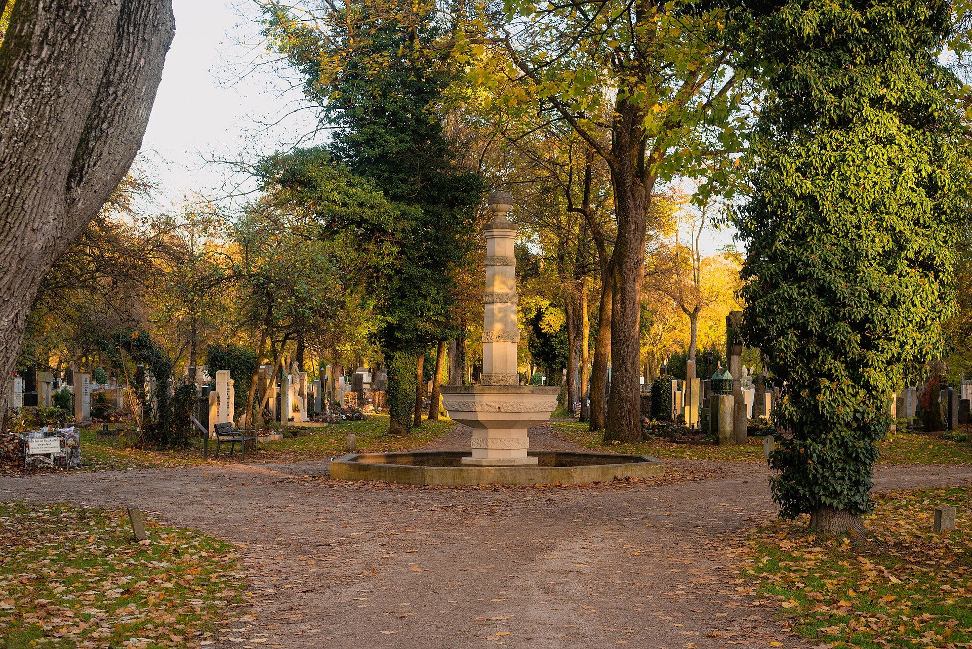 Steinerner Brunnen zwischen herbstlichen Bäumen und Grabstätten auf dem Münchner Westfriedhof